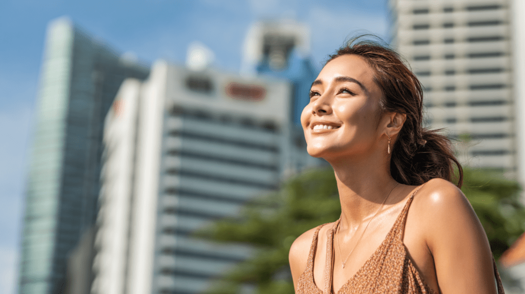 Woman smiling outdoors under the sun, showcasing radiant, sun-protected skin with a city backdrop, emphasizing the importance of sunscreen for skin protection in sunny environments.